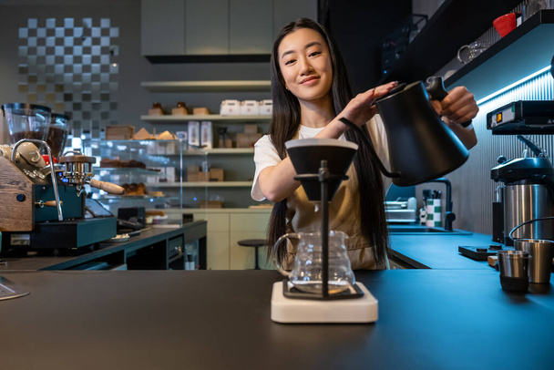 Waist-up portrait of a smiling barista tilting the kettle over the pour-over coffee maker funnel with filter - Фото, зображення