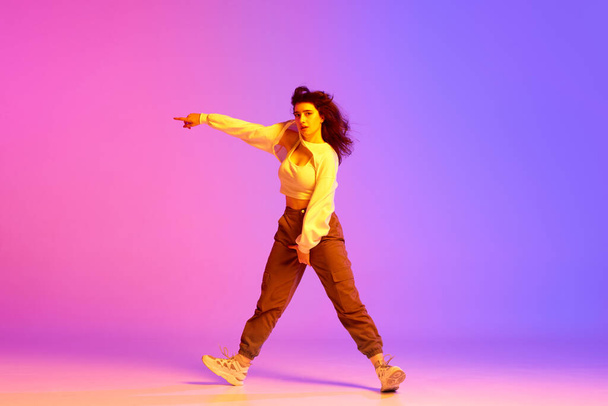 Dynamic image of young sportive girl, female hip-hop dancer training against gradient pink purple background in neon light. Concept of contemporary dance, youth, hobby, action and motion - Foto, Imagem