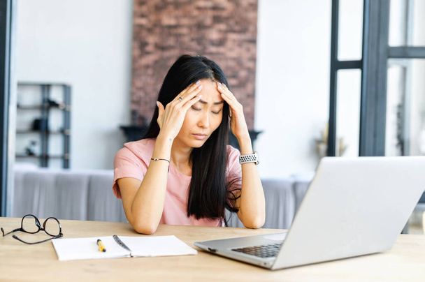 Tired woman sitting at the laptop after hard day, overworked female entrepreneur, wrinkled forehead, holding hand with the head, has a headache, a lot of information, glasses and notebook on the desk - Foto, imagen