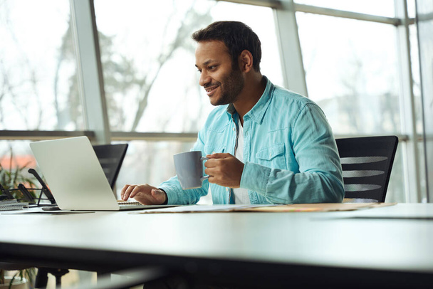 Szczęśliwy człowiek z kubkiem w ręku siedzi przy stole i pracuje na laptopie - Zdjęcie, obraz