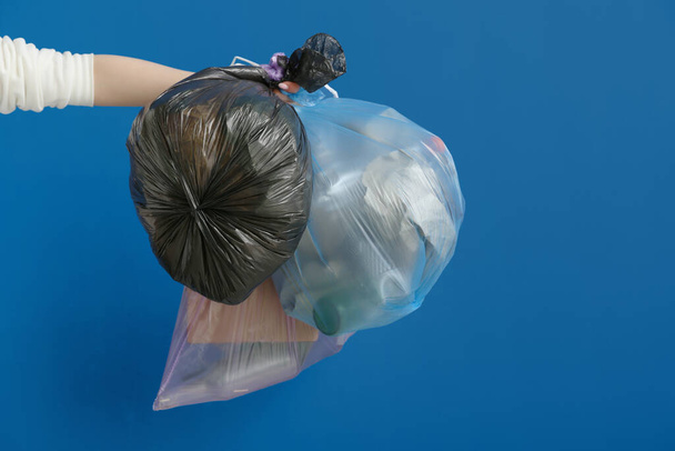 Γυναικείο χέρι που κρατά γεμάτες σακούλες σκουπιδιών σε μπλε φόντο - Φωτογραφία, εικόνα