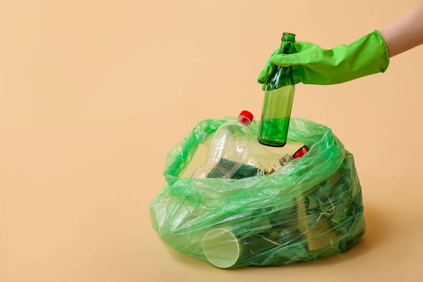 Female hand in rubber glove putting glass bottle into garbage bag on beige background - Foto, immagini