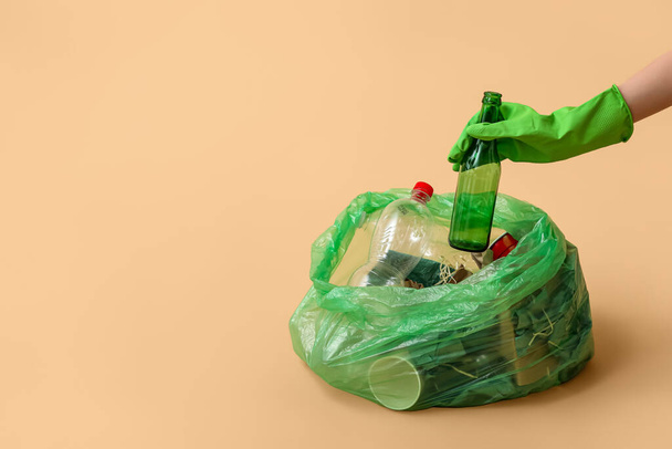 Lastik eldivenli bir kadın bej arka planda cam şişeyi çöp poşetine koyuyor. - Fotoğraf, Görsel