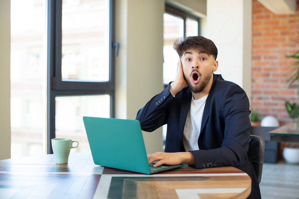 young handsome man feeling extremely shocked and surprised. freelance concept with laptop - Photo, image