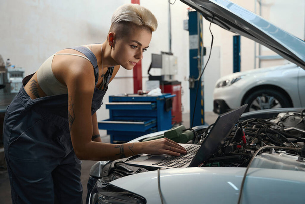 Female with a stylish haircut conducts computer diagnostics of a car, a woman is in a car repair shop - Photo, Image