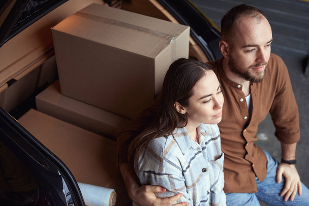 Vista superior de la foto del hombre tranquilo adulto abraza a una mujer mientras está sentado en el maletero de un coche cerca de cajas - Foto, imagen