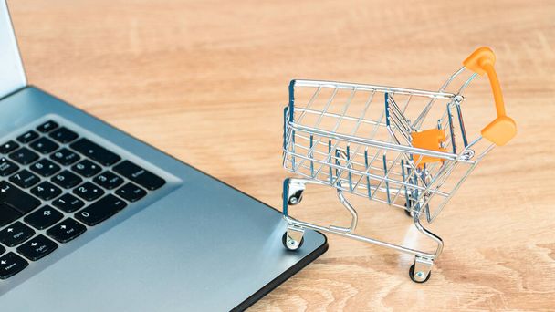 Online shopping concept. Shopping cart or trolley and laptop on wooden table - 写真・画像
