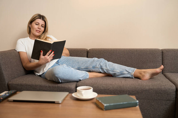 Charming female resting on comfortable sofa and enthusiastically reading book in living room of apartments - 写真・画像