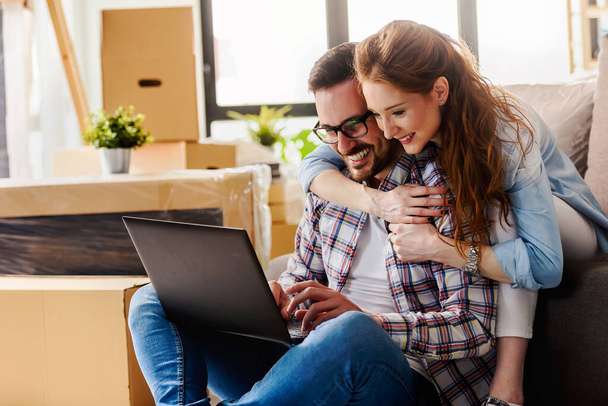 Young couple moving in new home. Sitting and relaxing after unpacking. Searching home decorating ideas on laptop - Photo, Image