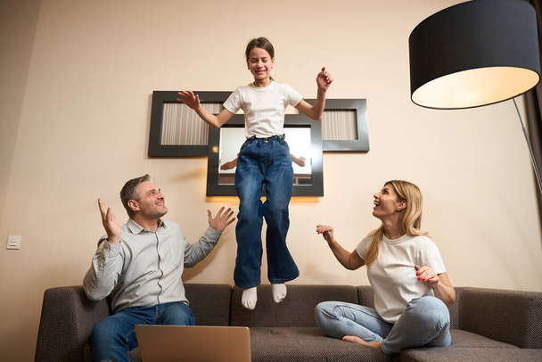 Pretty girl jumping on sofa while her smiling parents looking at her in modern room - Valokuva, kuva