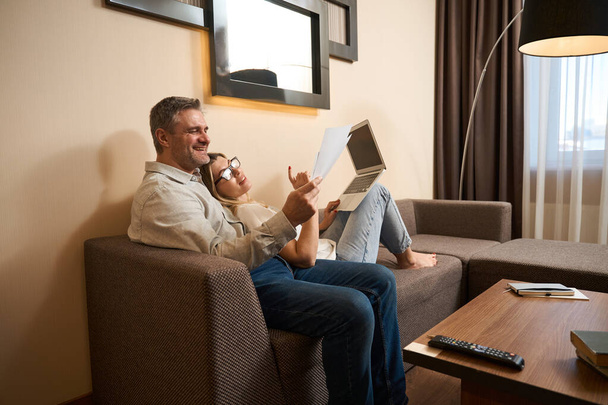 Handsome man showing paper sheet to smiling wife wearing glasses with laptop on her knees on sofa - Фото, изображение