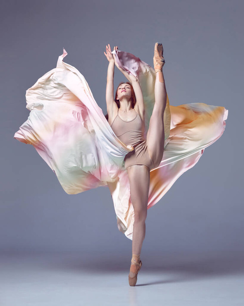 Ästhetik des klassischen Tanzes. Eine Grazien-Ballerina im Regenbogenkleid tanzt emotional mit Stoff über grauem Studiohintergrund. Konzept des klassischen Balletts, Inspiration, Schönheit, Tanz, Kreativität - Foto, Bild
