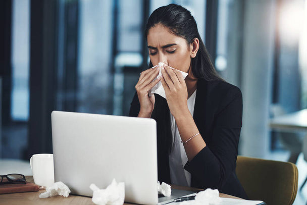 La gripe en el lugar de trabajo puede causar una reducción significativa de la productividad. una joven mujer de negocios sonándose la nariz mientras trabaja en una oficina - Foto, imagen