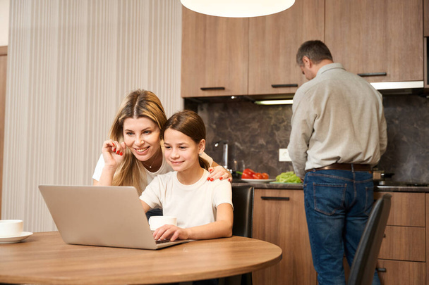 Little girl is sitting at the table with a laptop in the kitche and drinkind tea. Smiling woman is standing behind and huging daugter. In the background man is standing and preparing food - Foto, Imagem