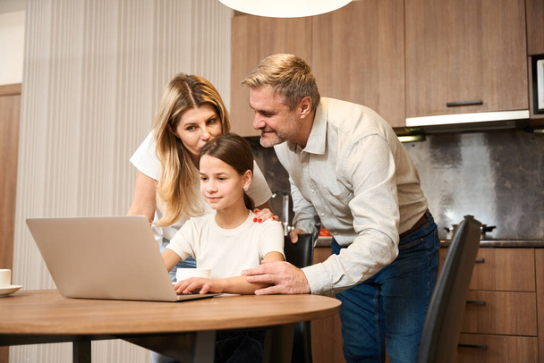 Little girl is sitting at table with laptop in the kitchen and drinking tea. Smiling woman is standing behind, kissing and huging her kid. Happy man is standing next to and holding the girl hand - Photo, Image