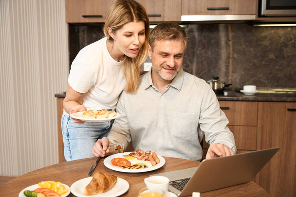 Smiling man is sitting at table in the kitchen. Beautiful lady is standing nearby and hugging man with one hand, in other hand she holding a plate of food. There are plates with dishes and laptop on - Foto, Bild
