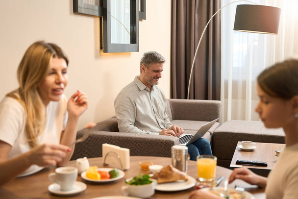 Woman and a little girl are sitting at the table and having breakfast and talking. Smiling man is sitting on sofa with laptop in hotel room - Фото, изображение