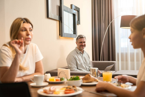Woman and a little girl are sitting at the table and having breakfast and talking. Smiling man is sitting on sofa with laptop in room - Photo, Image