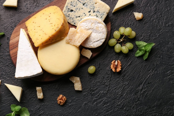 Ξύλινη σανίδα με διάφορους τύπους νόστιμου τυριού, ξηρών καρπών και σταφυλιών σε σκούρο φόντο - Φωτογραφία, εικόνα
