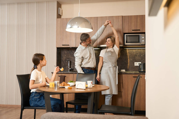 Little girl is sitting at the table, eating meal and drinking tea. Her parents and dancing in the kitchen in hotel room - Foto, Imagen