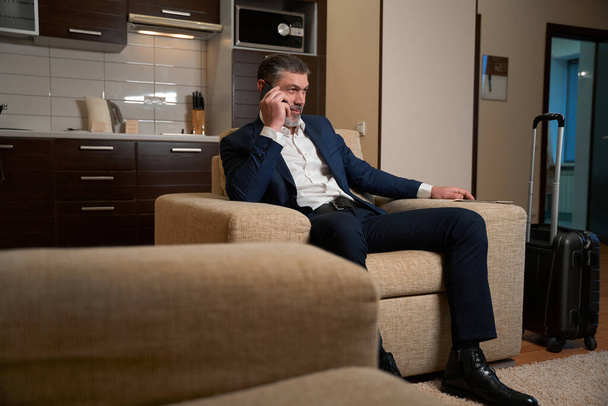 Erwachsener Mann im Businessanzug sitzt im Sessel und telefoniert im Hotelzimmer - Foto, Bild