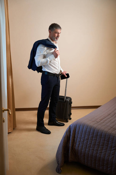 Дорослий чоловік в офісному одязі тримає ручку валізи в готелі
 - Фото, зображення