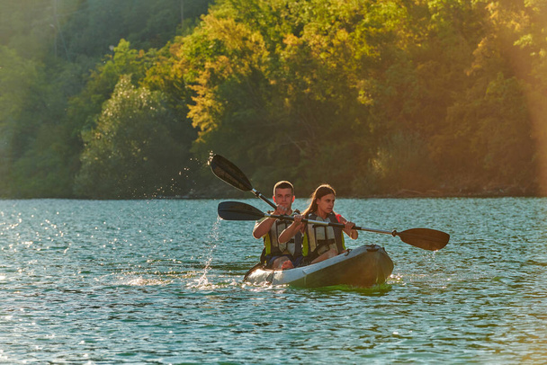 A young couple enjoying an idyllic kayak ride in the middle of a beautiful river surrounded by forest greenery in sunset time. - Zdjęcie, obraz