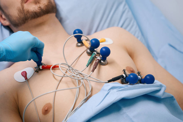 Cardiograaf zuignappen worden bevestigd aan een jonge patiënt, een man ligt op een ziekenhuisbed met een naakte romp - Foto, afbeelding