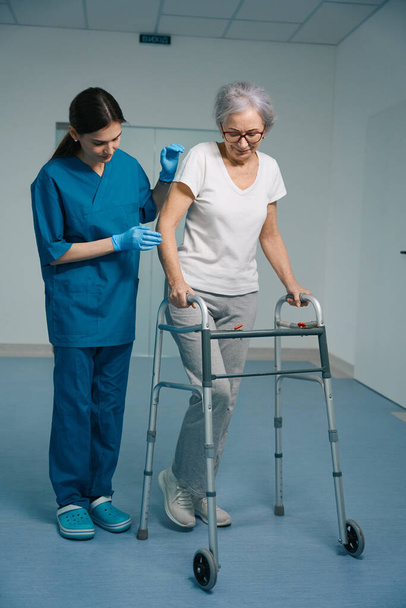 Young nurse accompanies an elderly lady, the patient moves with a walker - Фото, изображение