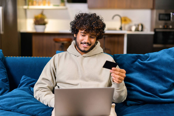 Happy young bearded Indian man 20s sitting on the sofa at home, using laptop pc computer and holding credit bank card, doing online shopping, ordering food delivery - Fotó, kép