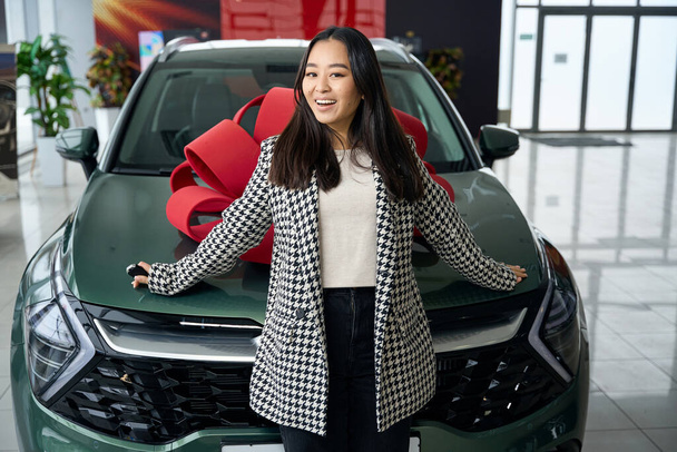 Jonge aziatische vrouw staat voor een auto met een rode strik, het vrouwtje heeft de autosleutels - Foto, afbeelding