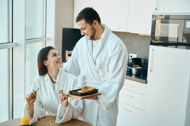 Loving young husband treats his charming wife to breakfast in a cozy atmosphere, the guys in comfortable bathrobes - Valokuva, kuva
