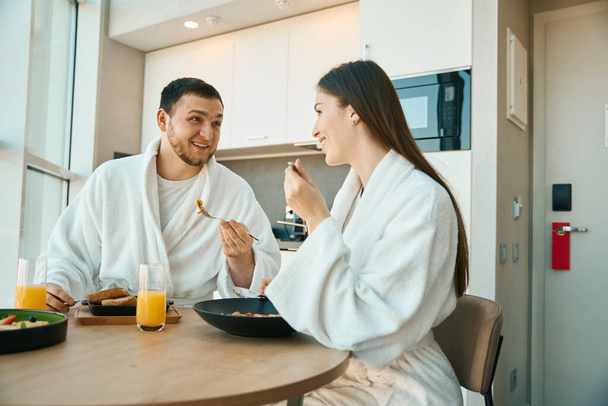 Счастливая молодая пара завтракает в уютной обстановке, парни в мягких халатах - Фото, изображение