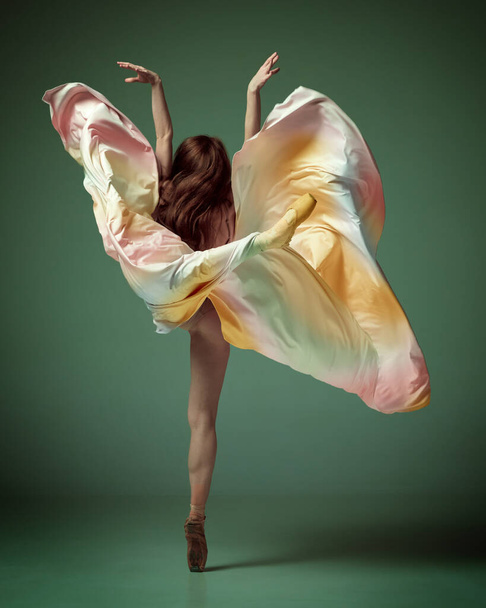 Estética de movimiento. Vista trasera de una encantadora bailarina, bailarina de ballet con un vestido colorido y elegante posando en puntillas sobre un fondo de estudio verde oscuro. Concepto de inspiración, danza, creatividad - Foto, Imagen