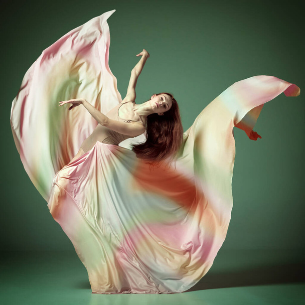 Portrait with one young girl, gorgeous ballerina wearing silk dress spinning in dance over dark green studio background. Concept of classical ballet, inspiration, beauty, dance, creativity, art - Foto, Imagen