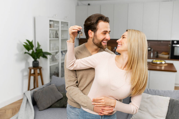 Happy young caucasian married couple holding key ring in hand, hugging and looking at each other with love, while standing in the modern kitchen of their new home. Excited owners of a family apartment - Fotó, kép