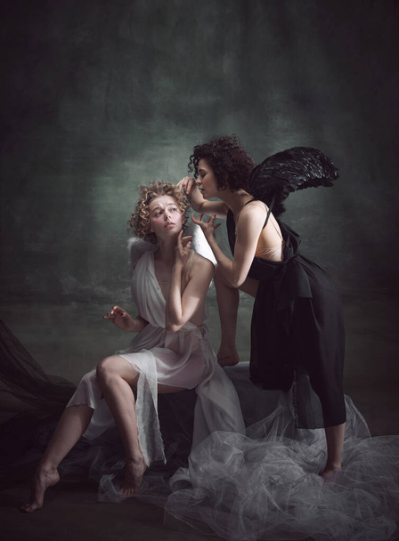 Devilish woman whispering to naive angel. Portrait of two women, angel and demon against dark, green, vintage background. Concept of history, remake, good and bad, creative photography - Fotoğraf, Görsel