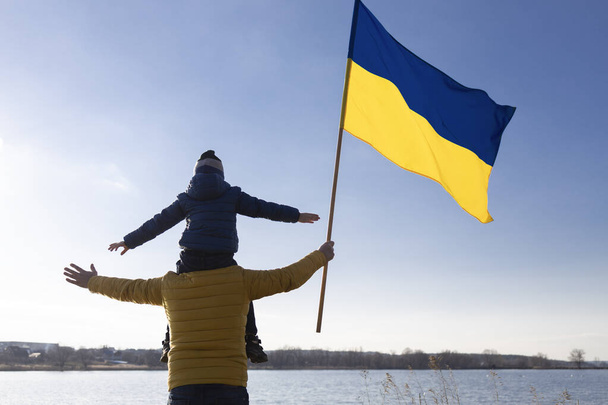Der unkenntliche Junge sitzt auf den Schultern eines Mannes, Vater mit Sohn, mit einer großen Fahne der Ukraine gegen den Himmel an einem sonnigen Tag. Patriotische Erziehung. Stolz, Siegesglauben. Schluss mit dem Krieg. Unabhängigkeitstag - Foto, Bild