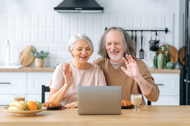 Happy caucasian pensioner couple, husband and wife, sit at home in the kitchen, use a laptop to communicate via online video conference with children and grandchildren, greet, smile joyfully - Foto, afbeelding