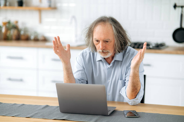 Frustrated caucasian gray haired elderly man, pensioner, financial mentor, sits at a table in the kitchen, looks confusedly at a laptop, is perplexed by the news or the result of work, gesturing hands - Zdjęcie, obraz
