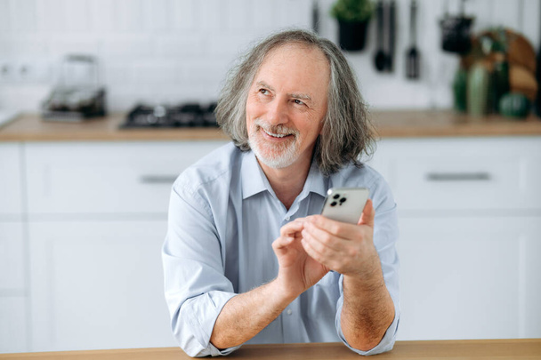 Positif satisfait homme aux cheveux gris caucasien mature avec barbe, assis à la maison dans la cuisine à la table, relaxant, en utilisant son smartphone, naviguer dans les médias sociaux, lire les nouvelles, faire du shopping en ligne, sourire - Photo, image