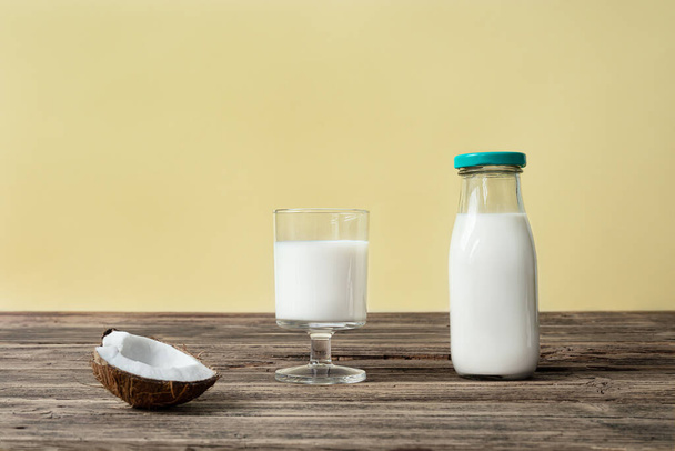 Bir bardak ve bir şişe vejetaryen hindistan cevizi sütü ve bir parça hindistan cevizi. Tahta masa, alternatif ve sağlıklı süt konsepti. - Fotoğraf, Görsel