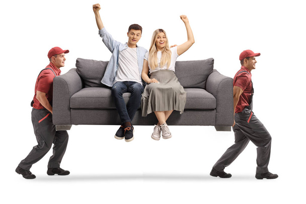 Movers carrying a young couple sitting on a gray sofa and gesturing happiness isolated on white background - 写真・画像