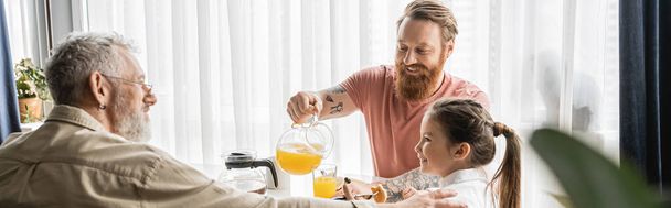 Smiling gay man pouring orange juice near daughter and partner at home, banner  - Foto, Bild