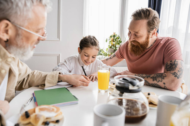 Smiling girl writing on notebook near gay dads and breakfast at home  - Foto, imagen