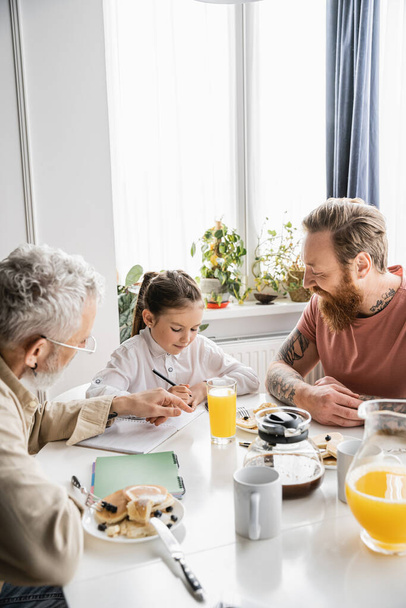 Homosexual fathers looking at daughter writing on notebook near breakfast and drinks at home  - Fotoğraf, Görsel