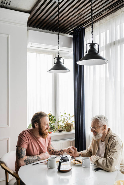 Tätowiertes gleichgeschlechtliches Paar hält Händchen bei Frühstück und Smartphone zu Hause  - Foto, Bild