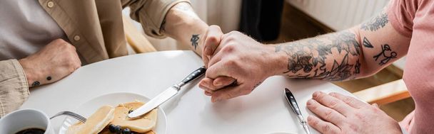 Vista recortada de pareja homosexual tatuada cogida de la mano cerca de café y panqueques en casa, pancarta  - Foto, Imagen