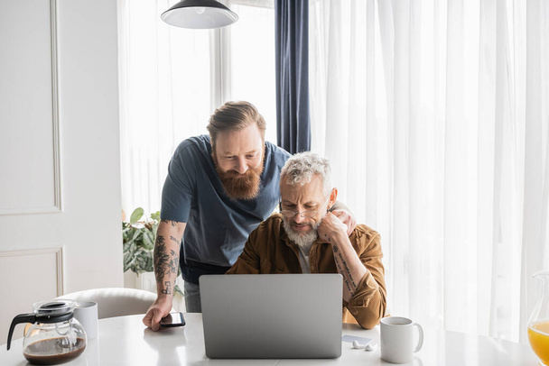 Tattooed gay man hugging mature partner using laptop near coffee at home  - Foto, immagini
