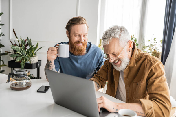 Smiling gay man holding cup of coffee near boyfriend during laptop at home  - Φωτογραφία, εικόνα